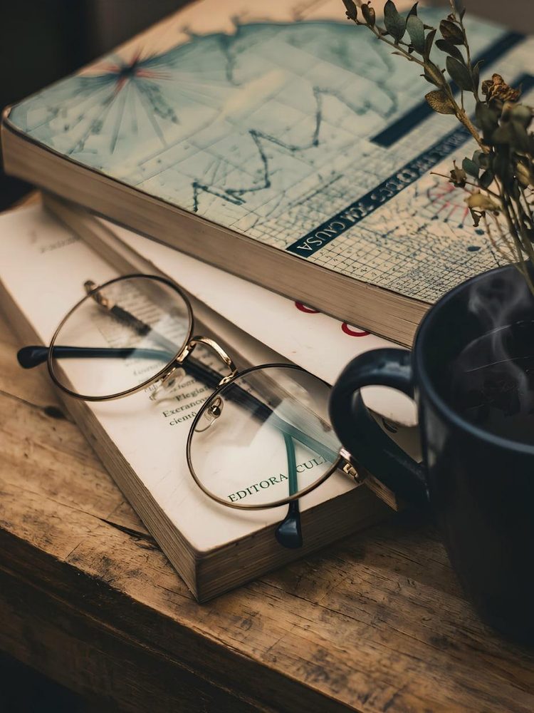 Reading glasses resting on a stack of books beside a map and a cup of coffee